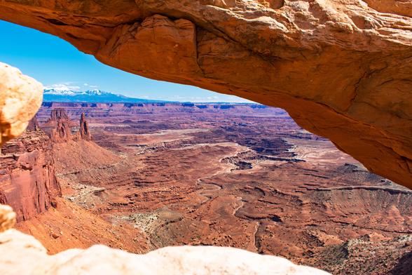 View through a natural red sandstone arch in a desert landscape, looking out over deep canyons, spires, and the distant La Sal Mountains under a clear blue sky.