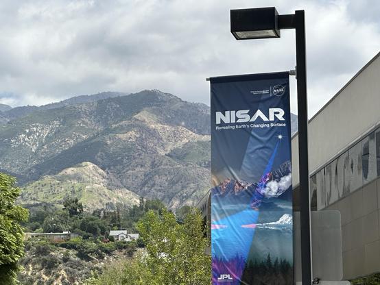 Photo of a banner hanging from a light pole. The banner has an artistic figure showing a beam of radar shining on Earth mountains, glaciers, lakes, and forests in shades of purple and pink. Text says NISAR Revealing Earth’s Changing Surface. Behind the pole is a metal and glass building and I the distance the San Gabriel Mountains.