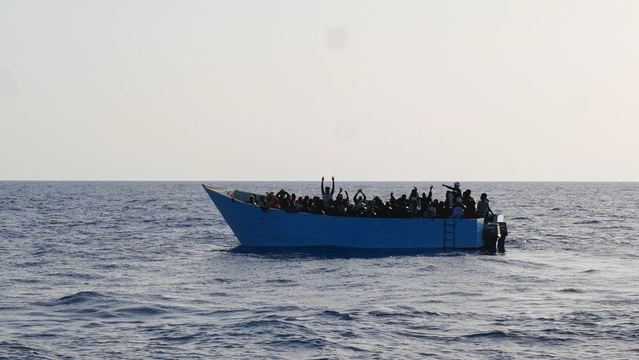Ein überfülltes blaues Holzboot, auf dem die Menschen dichtgedrängt zusammen stehen und sitzen. Einige winken mit erhobenen Armen. Niemand trägt eine Rettungsweste.