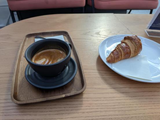 A Cup of Coffee on a wooden table next to it a Croissant on a white, porcelain plate