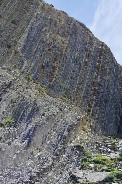 A steep cliff face features layered rock formations, showcasing geological striations and patches of vegetation. The sky above is partly cloudy.