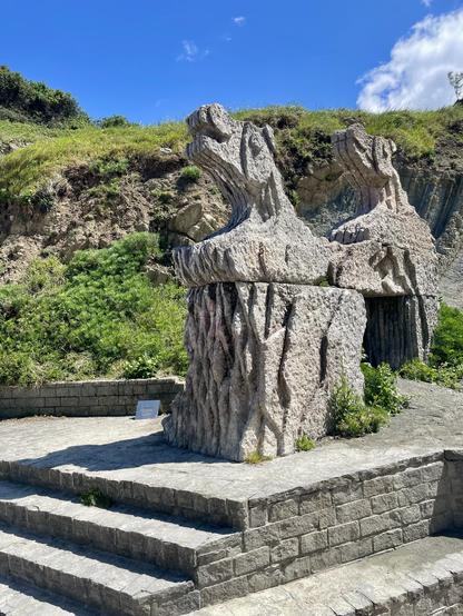 A large stone sculpture resembling two stylized animal heads stands on a stone platform. The background features grassy hills and a bright blue sky with a few clouds. Steps lead up to the sculpture, which is surrounded by lush greenery.