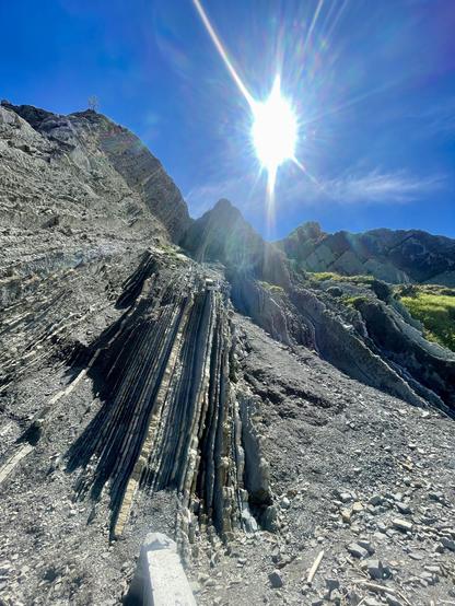 A steep, rocky hillside with layered geological formations under a bright sun against a clear blue sky.