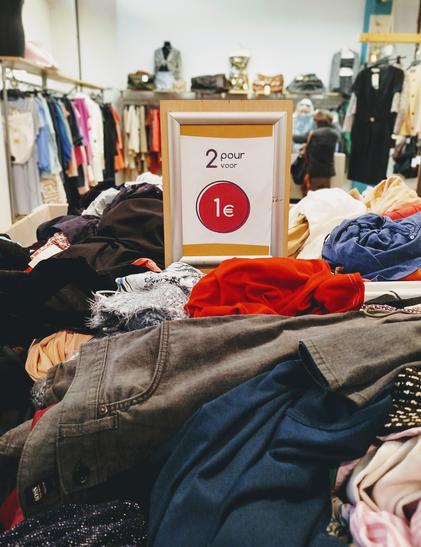 À l'avant plan, sur un tas de vêtements déposés dans un bac, une inscription indiquant "2 pour 1€".
On voit des tringles avec des vêtements à l'arrière plan.
