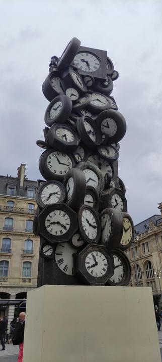 Photographie d'une sculpture en forme de colonne recouverte de dizaines d'horloges anciennes, toutes figées à des heures différentes. L’œuvre se dresse devant la gare Saint-Lazare à Paris, sous un ciel gris.