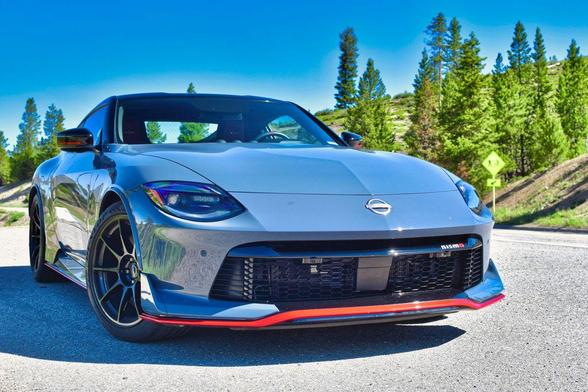 Front three-quarter view of a 2024 Nissan Z NISMO in metallic gray with red accents, parked on a mountain road with pine trees and blue sky in the background.