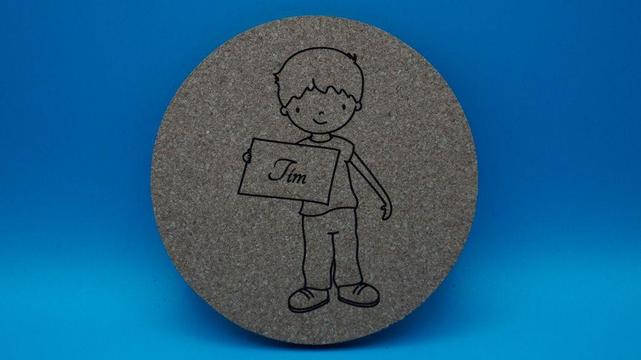 Runder Korkuntersetzer mit graviertem Motiv eines Jungen, der ein Schild mit dem Namen „Tim“ in den Händen hält.