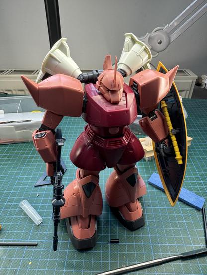The image features a large, articulated model robot with a predominantly maroon color scheme, wielding a shield in one hand and a rifle in the other. The robot is positioned on a green cutting mat, surrounded by various modeling tools and materials.