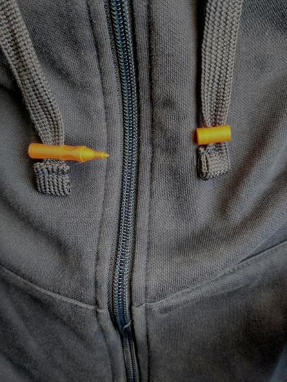 Links und rechts sind jeweils eine Kordel eined Hoody zu sehen. In der Mitte der Reißverschluss. Die linke Kordel hat einen orangenen Mini-Stift in Form eines Neuland Ouliner Stifts als Dekoration. Die rechte Kordel hat den zugehörigen Stiftdeckel in Miniatur-Format als Dekoration.