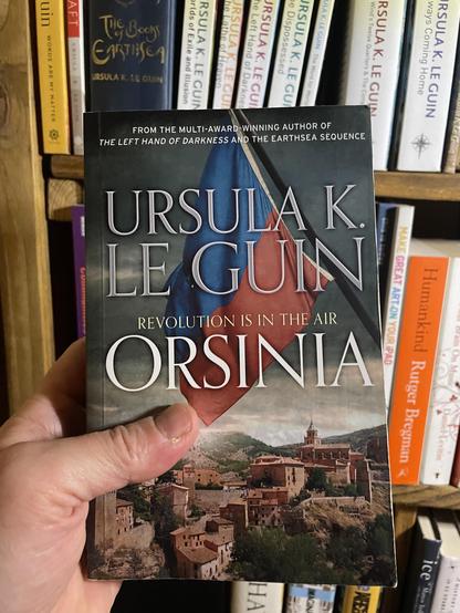 A white male hand holding up a copy of Ursula K. Le Guin's 'Orsinia' in front of a shelf of other Le Guin books.