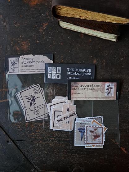 Three sticker packs on an old table top. First pack is called Botany sticker pack and it has four botanical illustration stickers. Second pack is called the forager sticker pack and it has seven illutrated stickers inspired by nordic nature cabin life. Third sticker pack has six stamp shaped stickers with painted mushroom illustrations.