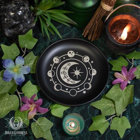 A "Moon Phase Metal Altar Dish" product is placed on a dark lace-covered surface. It's a shallow black-glazed metal bowl with moon phases and stars in a circle around a larger crescent moon and sun. Presented by Inked Goddess Creations.