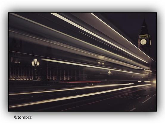 The photo captures a nighttime scene featuring Big Ben in London, taken using a long exposure technique.** This allows passing vehicles to leave dynamic light trails stretching across the width of the frame. The red and white lights create a sense of movement, contrasting with the still architecture of the city. In the background, the well-lit tower of Big Ben stands majestically, with its clock clearly visible. In the foreground, streetlights and sections of buildings add depth to the composition. The road displays the words "BUS LANE," slightly blurred due to the long exposure time.
Thanks to this technique, the photograph gains a dynamic quality, showcasing street traffic as striking streaks of light while the historic building remains a static focal point in the nighttime London scenery