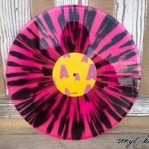 Eine farbenfrohe Schallplatte mit einem Tie-Dye-Muster aus Rosa und Schwarz, zentriert um ein gelbes Etikett mit lila Buchstaben. Der Hintergrund enthält Holztexturen.