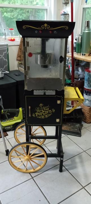 A dirty popcorn cart on a white tile floor. It is black with a yellow wheel and a yellow shelf on the right side. There are bins and tools in the background, in front of a window. There is a door to a storage bin on the cart that reads, "Old Fashioned Movie Time Pop Corn." 