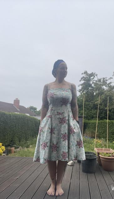 A person with their hair wrapped in dark blue cloth stands on a wooden deck on a drizzling day. They are wearing a light blue strappy dress with stylised red roses on it. Their hands are in the pockets. 