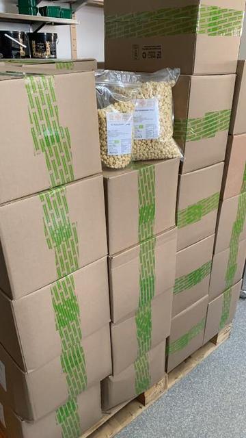 Eine Europalette auf der 5-6 Lagen graue Kartons mit grünem Klebestreifen gestapelt sind. Dazwischen zwei durchsichtige Beutel mit Bio Kakaobutter Chips.