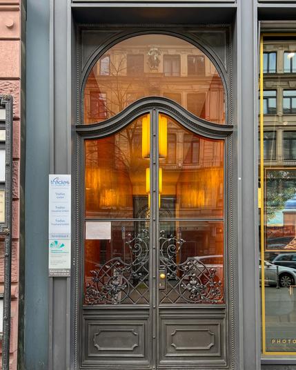 Grauer Eingang, fein zieselierte Windungen über Kassettenfläche hinterelgt mit Glas. Durch diese Eingagnstür sieht man die warm leuchtenden Lampen im Eingangsbereich sowie die Spiegelung Frankfurts
