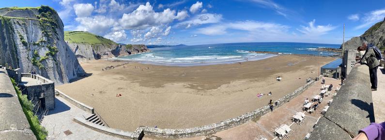 A panoramic view of a beach with golden sand and gentle waves, flanked by steep cliffs and greenery. People can be seen enjoying the sun, some walking along the shore. In the foreground, a stone pathway leads to a dining area with tables. 