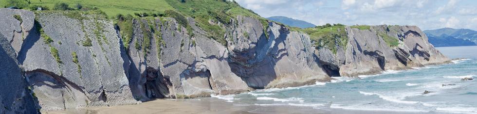 A panoramic view of rugged cliffs along a beach, with layered rock formations and greenery above. Waves gently crash onto the shore, and the sky is partly cloudy.
