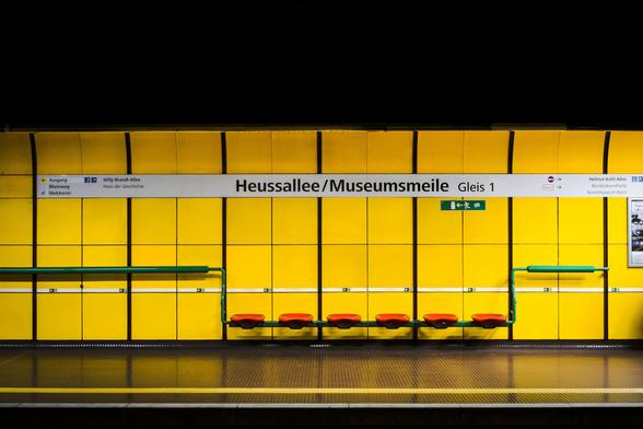 Das Bild zeigt den unterirdischen Seitenbahnsteig der U-Bahn-Station Heussallee/Museumsmeile in Bonn. Die Wände sind in einem kräftigen Gelb gehalten, im Kontrast dazu stehen die grünen Haltegriffe und die roten Sitzgelegenheiten.