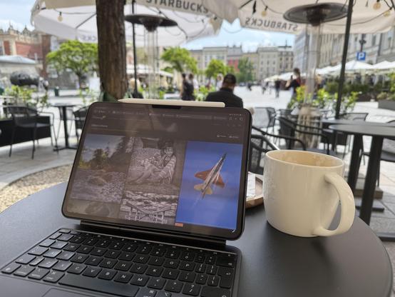 The photo shows an iPad with a keyboard on an outdoor café table at a Starbucks, located in a historic European city square -  Kraków, Poland. On the iPad screen, there’s a photo website open, displaying several artistic images. Next to the iPad is a large white Starbucks mug, partially filled with coffee. In the background, you can see cobblestone streets, people walking, historic buildings, trees, and large white umbrellas with “Starbucks” branding, suggesting a relaxed and scenic urban setting.