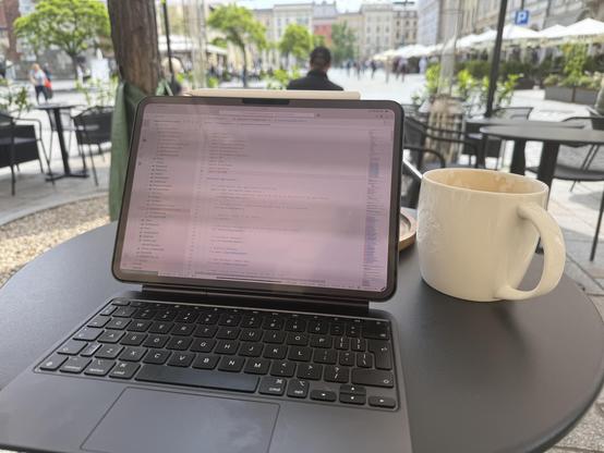 This photo is nearly identical in composition to the first one, but the screen of the iPad now shows a code editor (Visual Studio Code). Again, the coffee cup sits next to the device, and the same city square and outdoor café atmosphere is visible in the background, reinforcing the sense of combining work or hobby (programming) with leisure in a picturesque location.