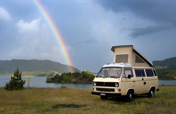 Au premier plan, à droite, un van VW beige, le toit ouvert.  Presque au centre, un arc-en-ciel semble rejoindre un petit îlot dans un lac de montagne. Le ciel est torturé, gris. En face, sur l’autre rive du lac, un rayon de soleil éclaire les sapins. 