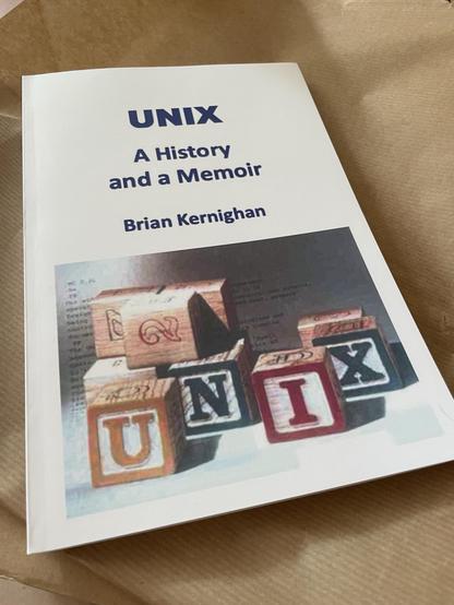 A book titled "UNIX: A History and a Memoir" by Brian Kernighan, featuring a cover with colorful blocks spelling "UNIX" and a background of text and designs. The book is resting on brown paper.