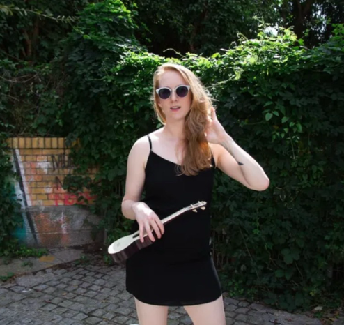 Sage stands in the sun, wearing sunglasses and a little black dress, holding a ukulele. 