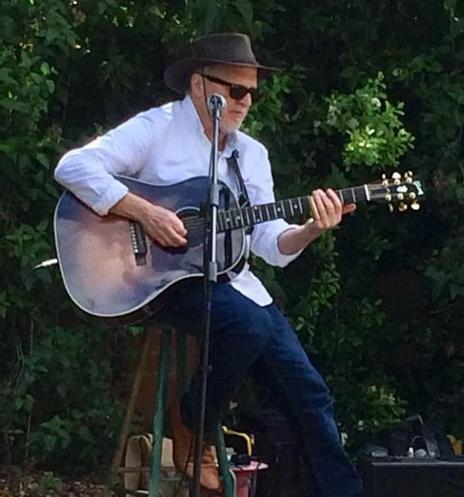 A man in a button-down shirt, wearing a hat and sunglasses, plays the guitar.