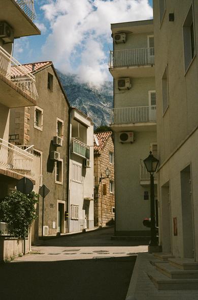 A street of Mediterranian houses, half in shadow, half in sunlight, with a tall mountain peaking out behind it, confidently grazing passing by clouds.