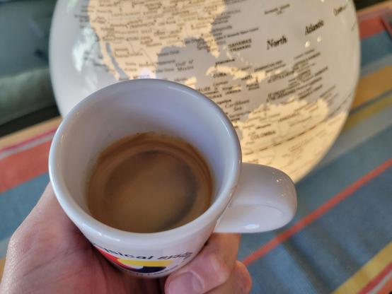 A white, 3 ounce, ceramic espresso (demitasse) cup with a shot of espresso, held in a hand in front of an illuminated globe centered on the Gulf of Mexico, which rests on a multi-colored (rust, light-green, red, light-gray, and blue), striped table runner. The blue text on the cup reads "Cyber-Physical Systems Research Center" and wraps in a circle around the logo, which looks like a loosely interlaced, rounded triquetra with thick knots in solid red, yellow, and blue (each its own color). 
