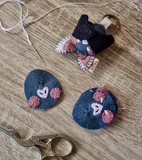 Picture or three bat felt keychains more or less finished set on a desk. 
The one on top is fully done and has its wings closed. The one on the bottom right has the eyes embroidered, but no wings or ears, and the one at the bottom doesn’t even have eyes yet.