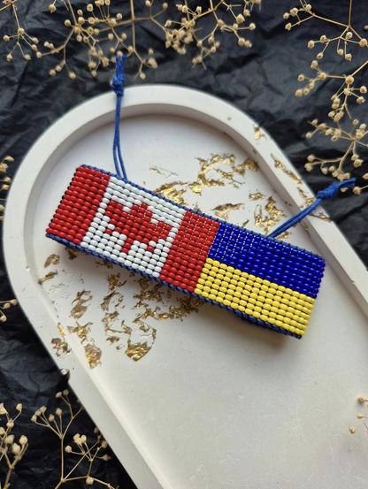 The photo shows a handmade beaded bracelet, half of the bracelet is the flag of Canada, the other half is the flag of Ukraine.