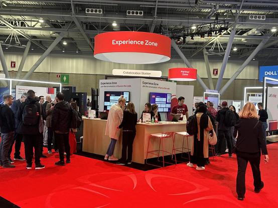 interior of a tech conference with 20-30 people, red carpet beneath and warehouse ceilings. The banner above this particular booth says experience zone. 