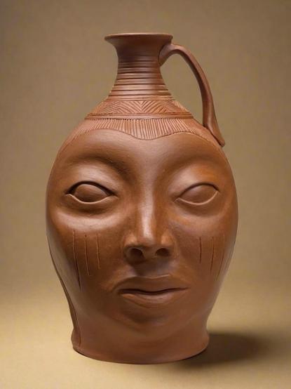 A ceramic jug made by Osa Atoe, sculpted to resemble a human face, featuring detailed facial features, expressive eyes, and decorative markings. The jug has a narrow neck and a handle, set against a neutral background. It has elements of a Southern face jug and the Ife terracotta heads combined. The color of the piece is a dark reddish brown, derived from wild clay terra sigillata. 