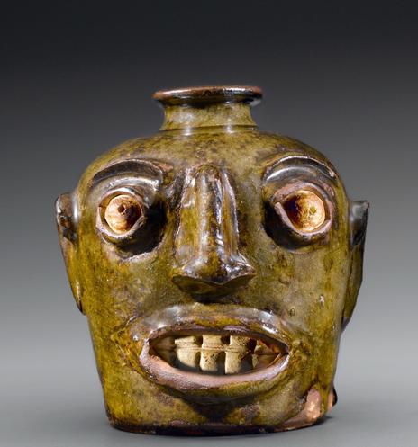 A ceramic vessel shaped like a human face, featuring exaggerated facial features, large eyes, and a wide mouth displaying teeth. The surface has a greenish-brown glaze, giving it an aged appearance. The jug is from Edgefield, South Carolina and was created by an unknown enslaved potter. 