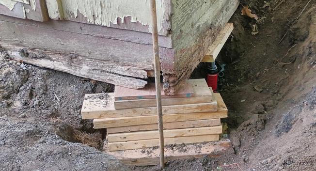 The corner of a shed has a bunch of blocking supporting it, and a little red 4ton hydraulic jack supports the more solid edge. This is all below ground level. 
The lower part of the wood is rotting away.