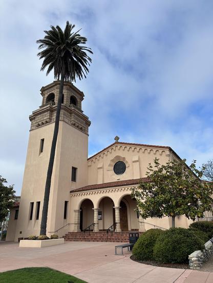 An architectural view of a building featuring a prominent bell tower and a palm tree. The structure has a warm-toned exterior with decorative arches and a tiled roof, surrounded by landscaped greenery and a paved area. The sky is partly cloudy.