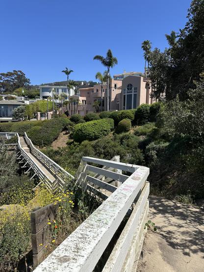 A scenic view featuring a white wooden bridge leading to a lush, green area with flowering plants. In the background, there are various coastal-style homes, including a prominent pink house, under a clear blue sky.