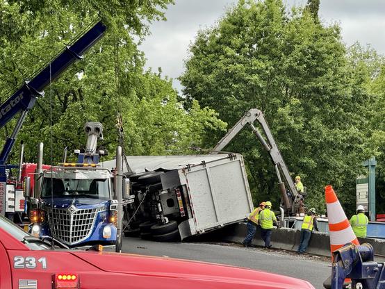 An overturned truck is being lifted by a tow truck on a roadside, with several workers in safety vests assisting. Trees are visible in the background, and a traffic cone is in the foreground.