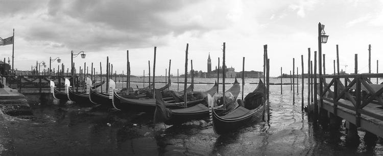 Panorama in black and white of a row of gondolas bobbing in the lagoon, the silhouette of a bell tower across the water.