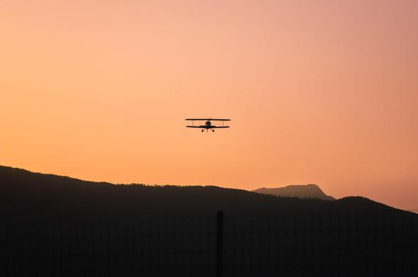 A silhouetted biplane flying against a gradient orange and pink sunset sky with dark rolling foothills below; faint fence lines are visible in the foreground.