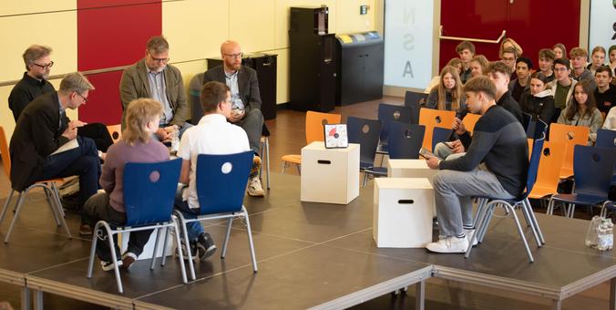 Foto einer Podiumsdiskussion in der Aula einer Schule. Auf dem Bild sind Schülerinnen und Schüler zu sehen, die Experten aus verschiedenen Bereichen zu Fragen rund um Geschichte, Medien und Politik befragen.