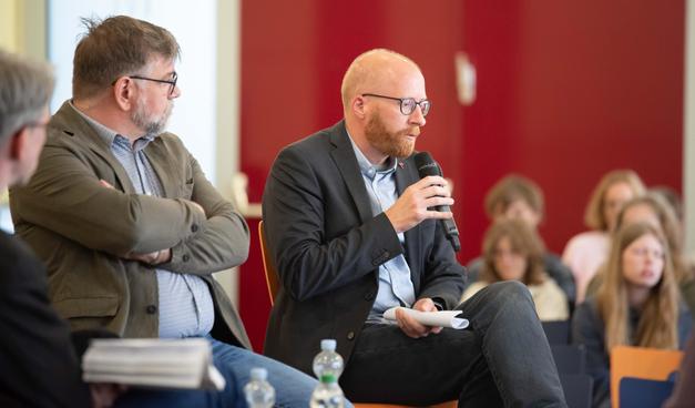 Foto einer Podiumsdiskussion in der Aula einer Schule. Auf dem Bild sind Schülerinnen und Schüler zu sehen, die Experten aus verschiedenen Bereichen zu Fragen rund um Geschichte, Medien und Politik befragen.