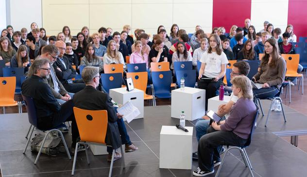 Foto einer Podiumsdiskussion in der Aula einer Schule. Auf dem Bild sind Schülerinnen und Schüler zu sehen, die Experten aus verschiedenen Bereichen zu Fragen rund um Geschichte, Medien und Politik befragen.