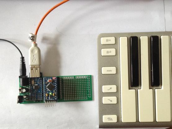 Photo showing the described DIY PCB in use.  It is a rectangular PCB 100x35 mm.  An Arduino Pro Mini and mini USB Host shield are mounted on the board.  The board has power from a barrel jack and the photo shows a USB MIDI keyboard plugged into the USB socket on the mini USB Host shield.
