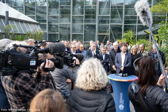 Angehörige der Universität Jena und Gäste stehen im Innenhof des Botanischen Gartens und warten auf einen Laptop schauend auf die Verkündung der künftig geförderten Exzellenzcluster. Medienvertreter*innen filmen das Geschehen.