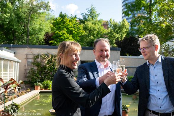 Prof. Dr. Kirsten Küsel, Sprecherin des Exzellenzclusters „Balance of the Microverse“, Prof. Dr. Andreas Marx, Präsident der Universität Jena und Prof. Dr. Johannes Grave, Sprecher des Exzellenzclusters „Imaginamics“, stoßen im Innenhof des Botanischen Gartens mit Sektgläsern an (v.l.n.r.).